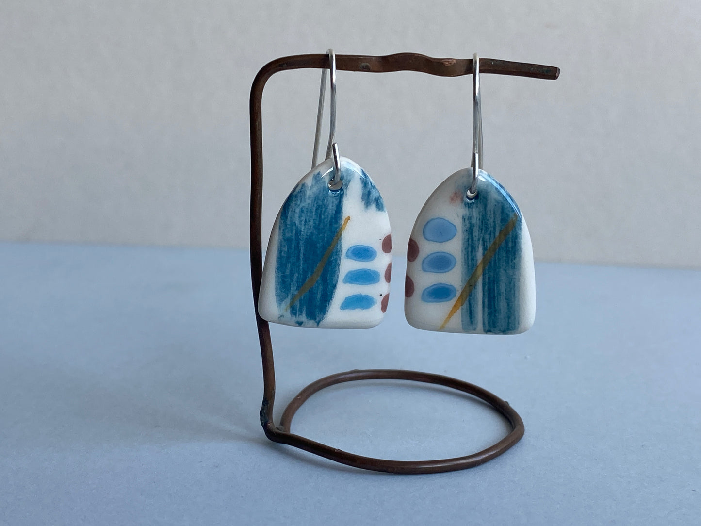 A pair of handmade ceramic earrings with a coastal vibe, featuring blue, white, and hints of orange patterns, hanging from sterling silver wires.
